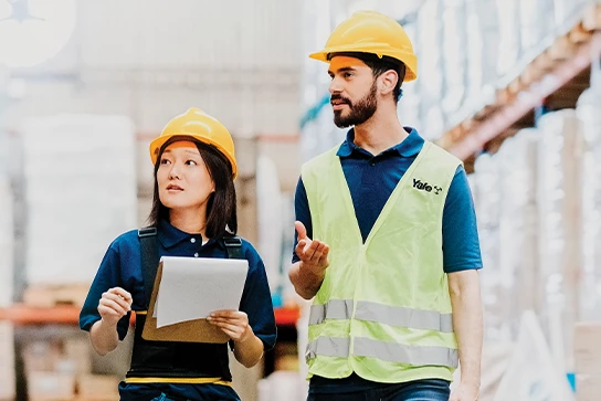 Two Yale warehouse workers with hard hats on walking down an aisle, with one holding a clipboard