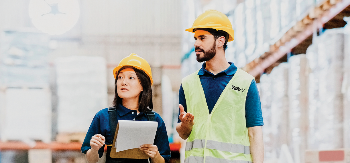 Two warehouse workers with hard hats on walking down an aisle, with one holding a clipboard