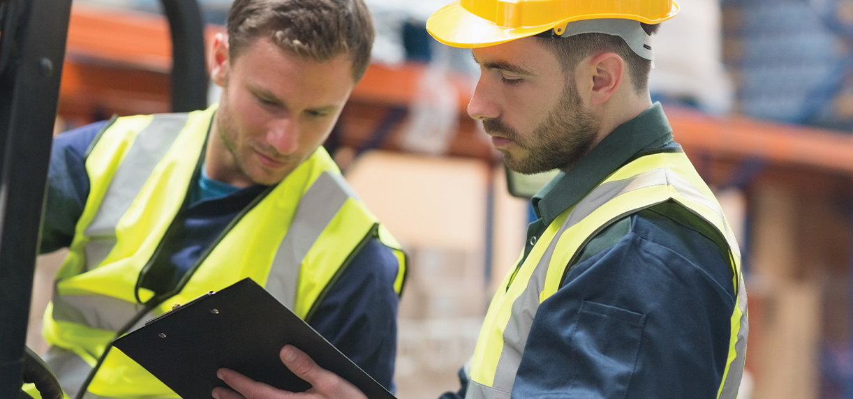 Two warehouse workers, one wearing a hard hat look at a clipboard in hand